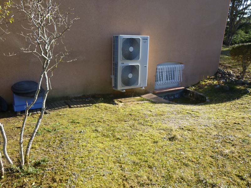 Unité extérieure Toshiba de climatisation double sur mur brun, près d'un arbre nu, d'une entrée souterraine grillagée et d'une pelouse verte.