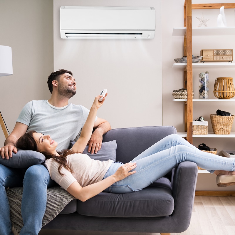 Couple détendu sur un canapé, la femme réglant le climatiseur mural avec une télécommande. Scène de confort estival.