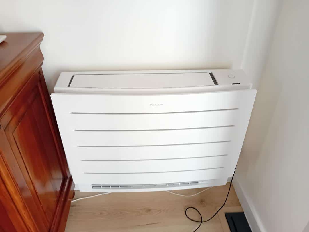 Climatiseur Daikin blanc, unité basse à lamelles près d'un meuble en bois. Logo Daikin visible sur l'appareil moderne.