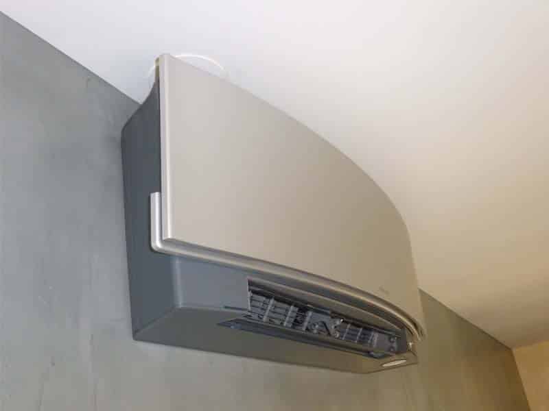 Climatiseur mural bi-ton gris, avec panneau supérieur incurvé, installé sur un mur texturé gris sous un plafond blanc. Sa sortie d'air est visible.