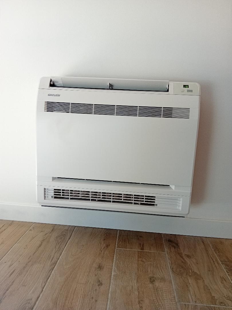 Climatiseur Sinclair blanc posé au sol contre un mur blanc, affichant '19' sur son écran. Un parquet en bois clair est visible.