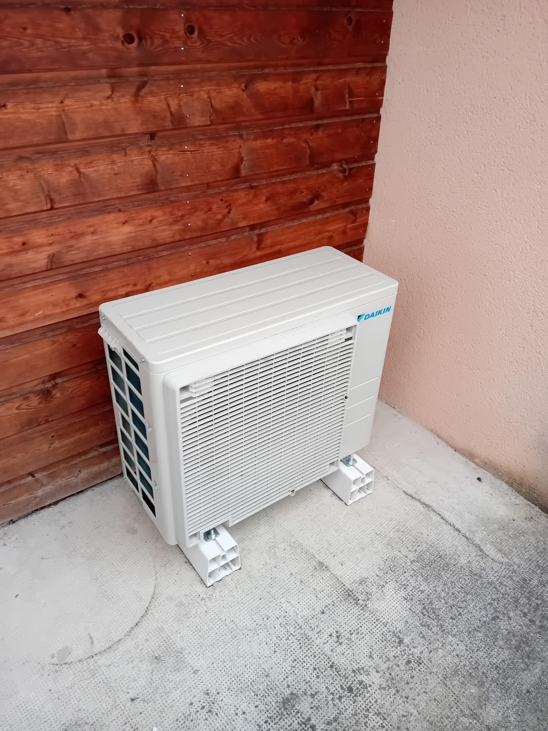 Unité extérieure de climatisation Daikin blanche sur supports, adossée à un mur en bois et un mur en stuc rose pâle, sol en béton texturé.