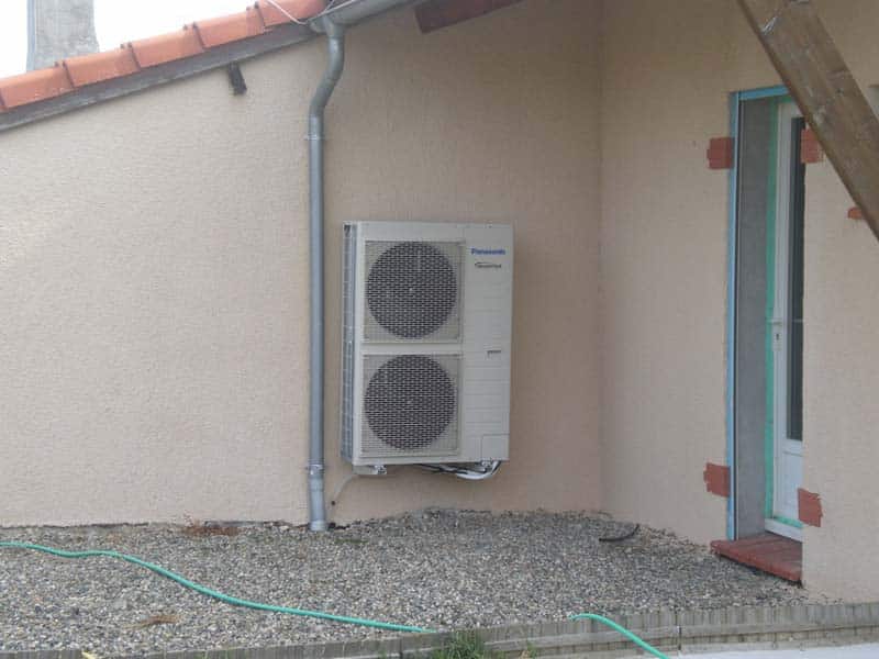 Unité extérieure Panasonic Inverter de pompe à chaleur sur un mur en crépi beige avec gouttière, porte et gravier au sol.