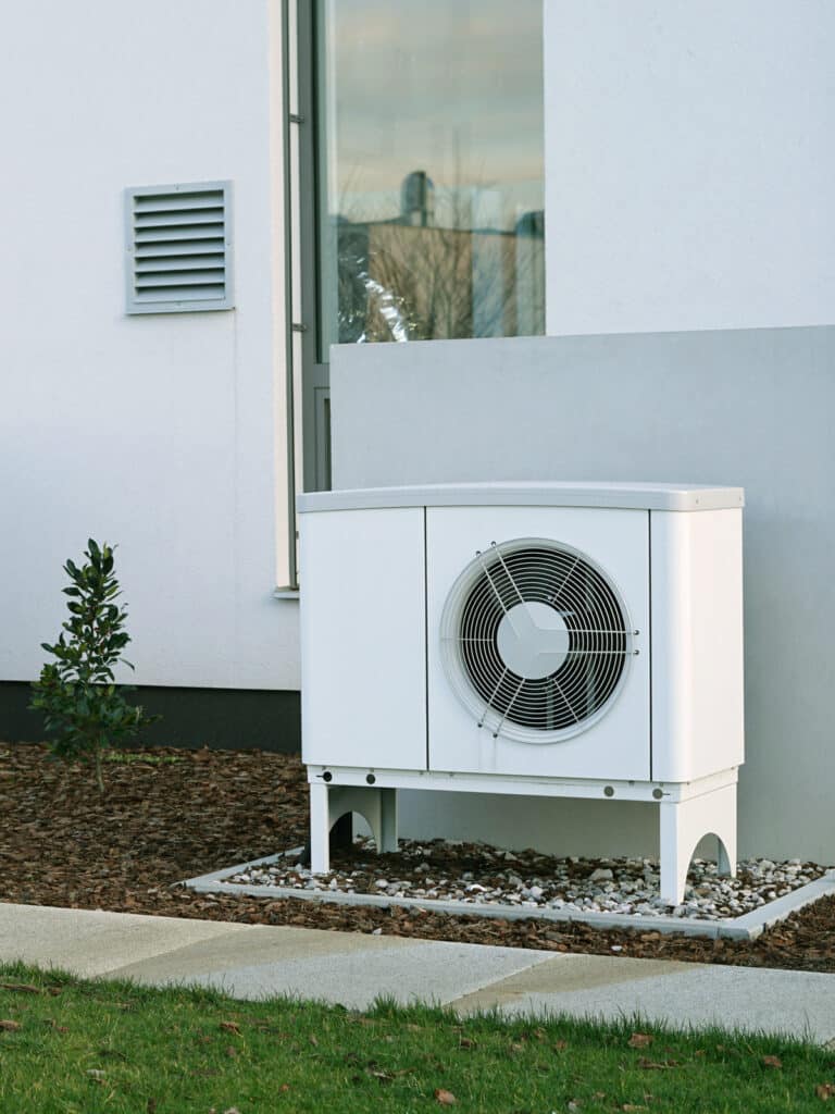 Pompe à chaleur extérieure blanche avec grille de ventilateur, installée sur des galets près d'une maison moderne et d'un arbuste.