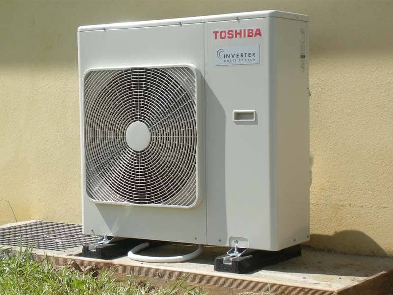 Unité extérieure Toshiba Inverter Multi System sur supports au sol, avec grille de ventilateur ronde, adossée à un mur jaune.